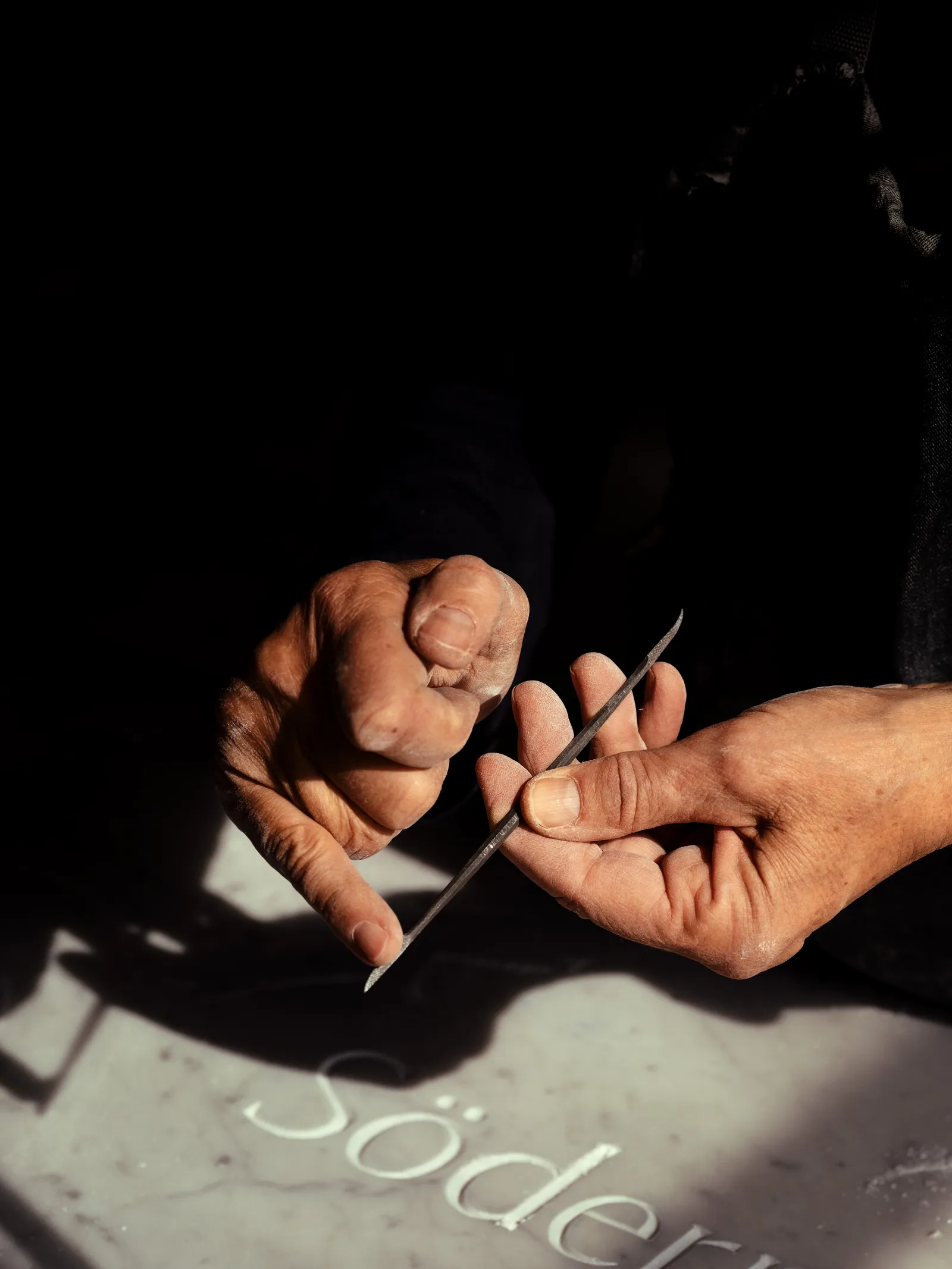 A carver’s hands gripping a mallet and chisel, engraving lettering into a marble slab in raking sunlight.