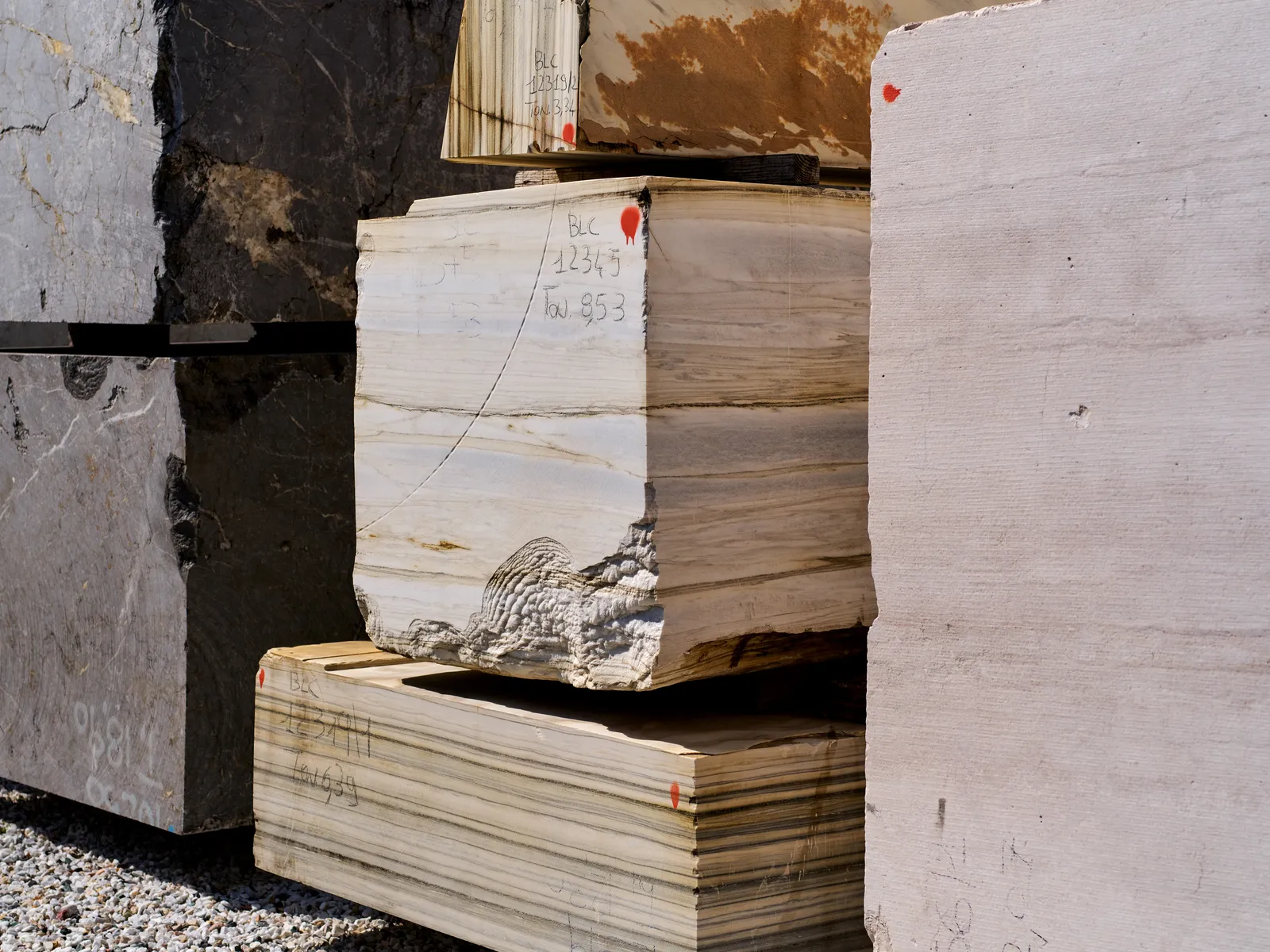 Marble blocks stacked in a stone yard, each marked with handwritten grading codes and a red dot.