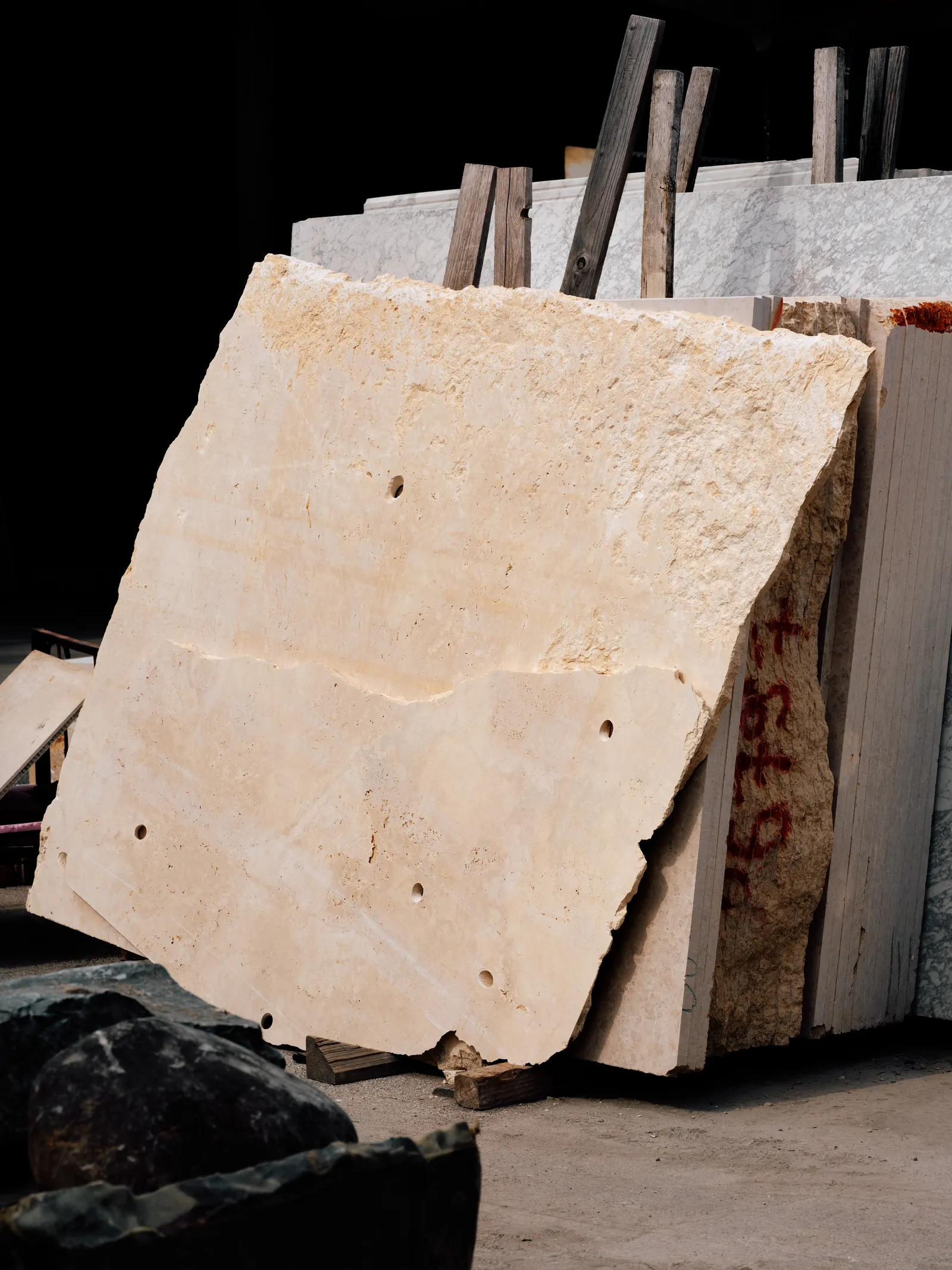 A roughly cut travertine slab, pale and porous, leaning against stacked marble in a dim stone yard.