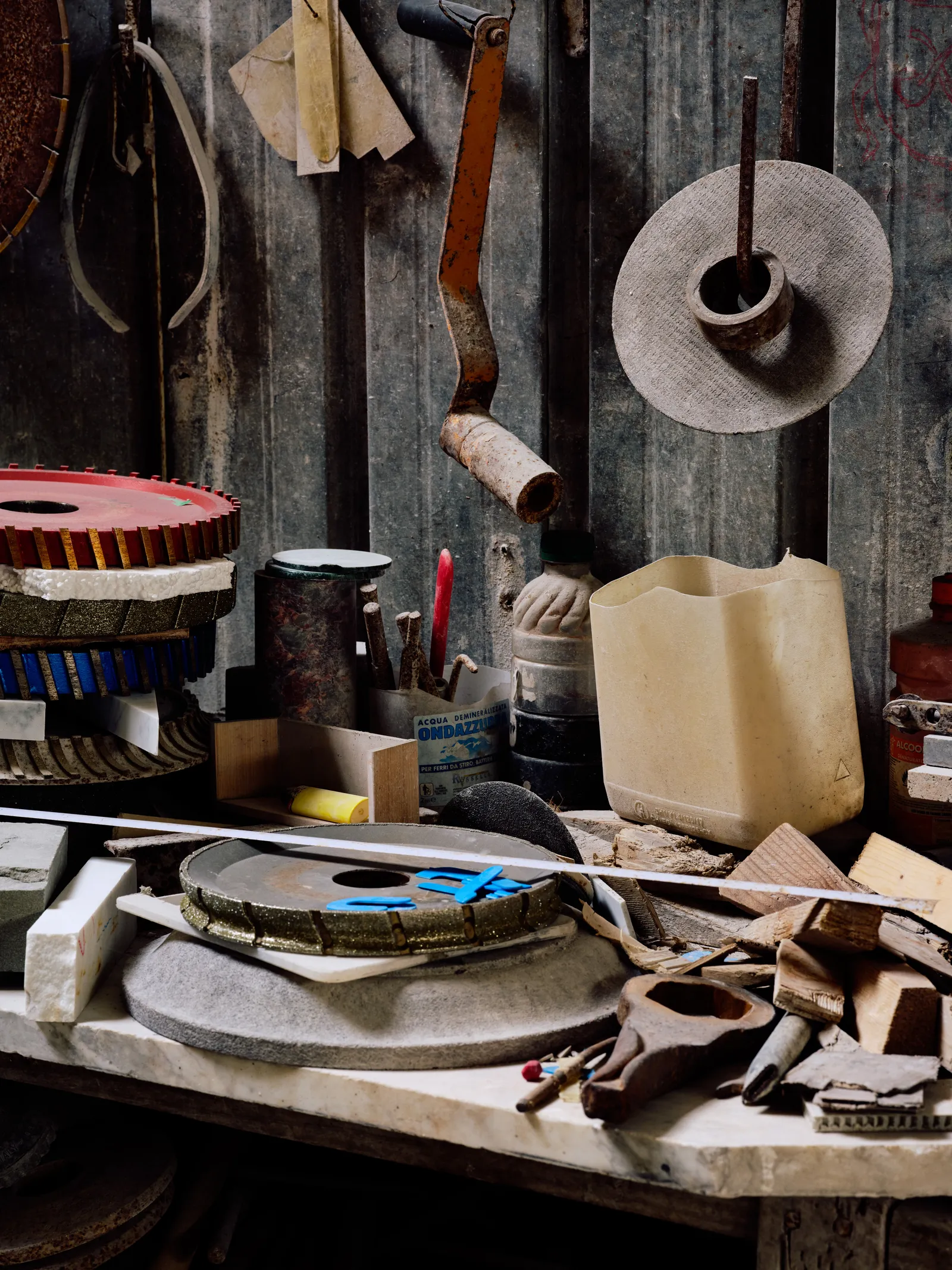 A cluttered stone-carving workbench with stacked grinding discs, hand tools, and a tape measure, sanding pads hanging on the wall behind.