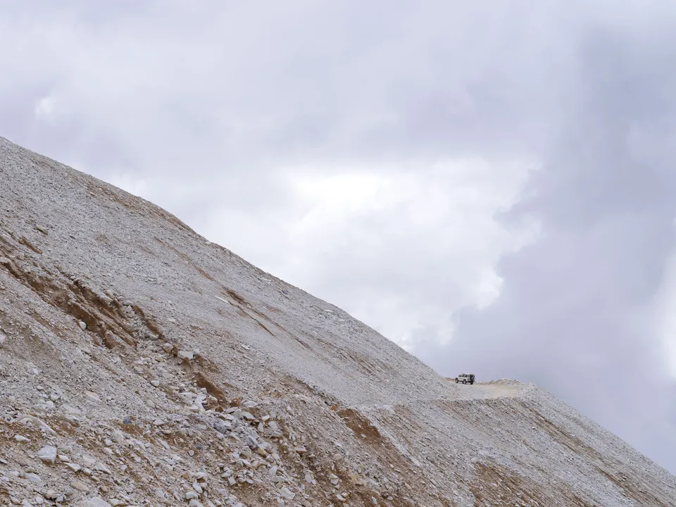 A pale quarry hillside in the Carrara mountains, a small vehicle barely visible on the ridge road.