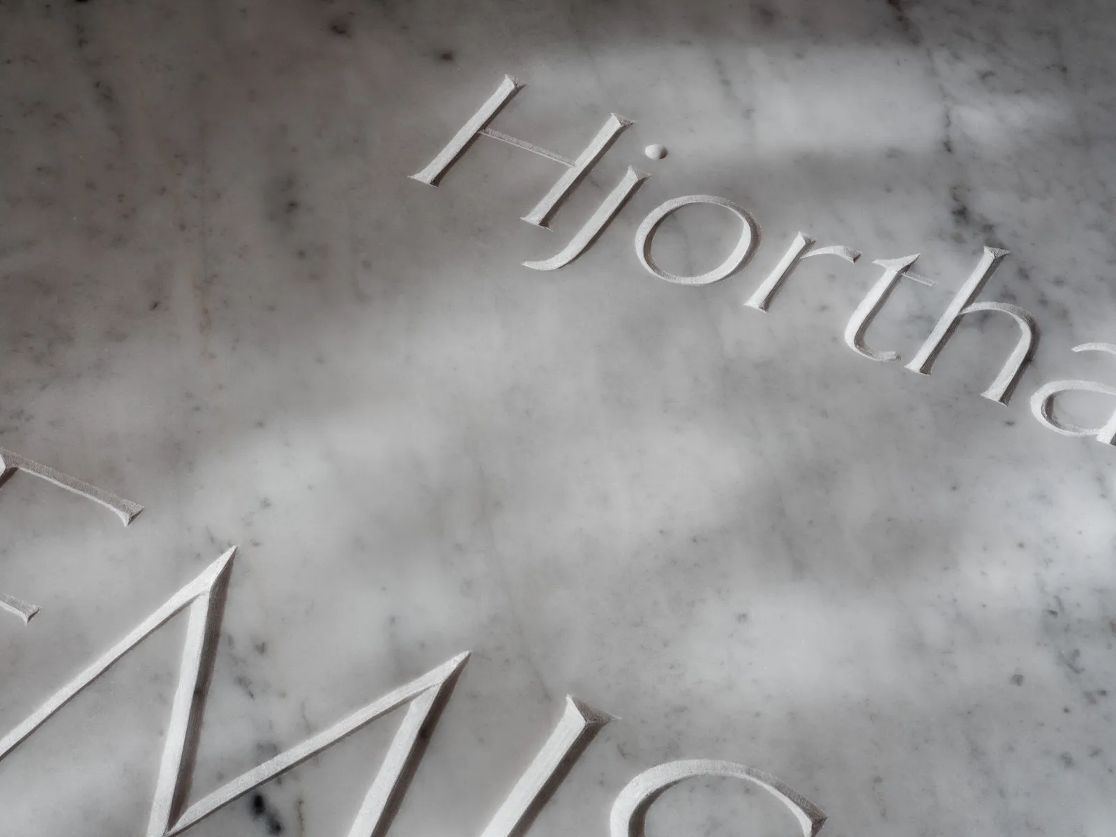 Extreme close-up of Hjorthagen carved into marble, raking light casting shadows across the letterforms.